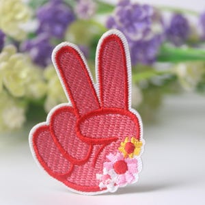 May include: A pink peace sign with white outline and embroidered flowers. The flowers are pink, white, and yellow.