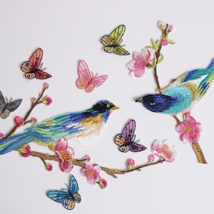 May include: Embroidered patches featuring colorful birds and butterflies perched on flowering branches. The birds have blue, green, and yellow plumage, while the butterflies are in shades of pink, orange, and green. The patches are set against a white background.