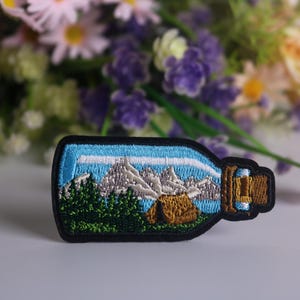 May include: An embroidered patch showing a glass bottle containing a mountain scene. The bottle is blue and white, with a brown cork. The mountain scene is green and white, with a small brown tent in the foreground.
