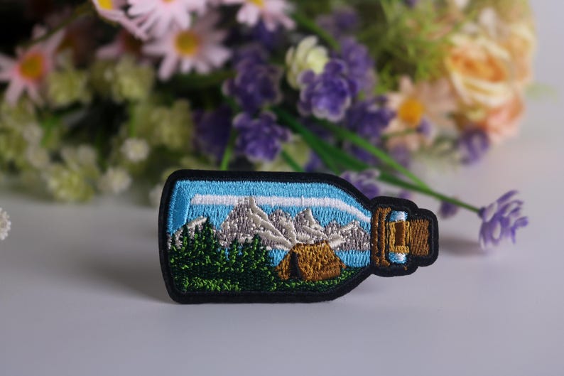 May include: An embroidered patch showing a glass bottle containing a mountain scene. The bottle is blue and white, with a brown cork. The mountain scene is green and white, with a small brown tent in the foreground.