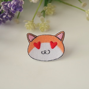 Puede incluir: Un parche bordado blanco y naranja de un gato con corazones rojos por ojos. El gato tiene una sonrisa simple.