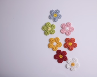 May include: Assortment of crocheted flower embellishments in various colors, including light blue, green, yellow, orange, red, and white. The flowers are arranged on a white background.