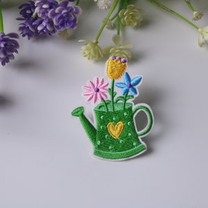 Op de afbeelding: Groene gieter met een hartvormig ontwerp en drie geborduurde bloemen in roze, blauw en geel.