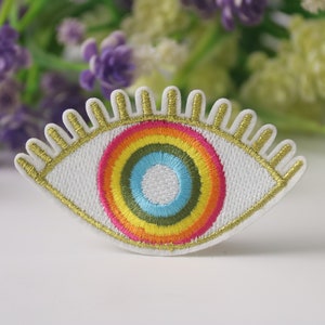 May include: A white embroidered eye patch with a rainbow iris and gold eyelashes. The patch is perfect for adding a whimsical touch to clothing, bags, or other accessories.