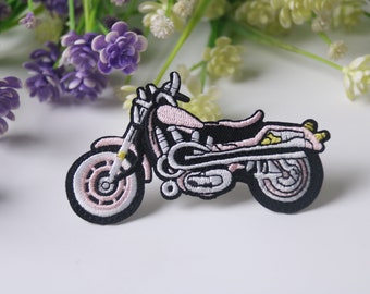 Puede incluir: Parche bordado con un diseño de motocicleta rosa y negra. El parche tiene un contorno detallado con detalles en blanco y amarillo. La motocicleta está sobre un fondo blanco con flores moradas y blancas.