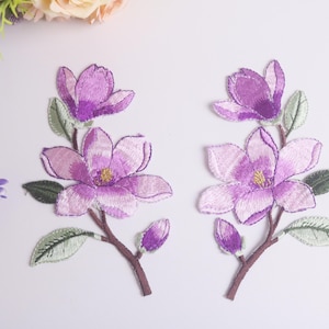 May include: Two embroidered floral patches featuring purple magnolia blossoms with green leaves and brown stems. The patches have a detailed, textured appearance, perfect for fabric embellishment or crafting projects.