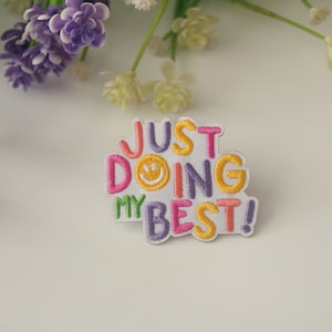 May include: A white embroidered patch with the text "JUST DOING MY BEST!" in colorful letters. A yellow smiley face is included in the word "DOING".