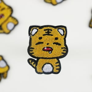Puede incluir: Un parche de hierro amarillo bordado de un tigre de dibujos animados con contornos negros. El tigre tiene una boca ancha abierta con una lengua roja que sobresale.