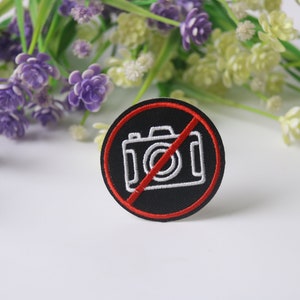 Puede incluir: Un parche bordado negro y rojo con un icono de cámara blanco tachado. El parche es una señal de prohibición de fotografía.