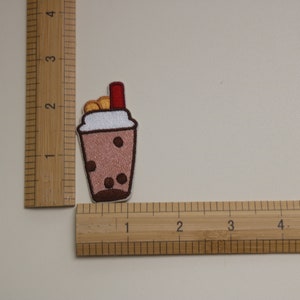 Może przedstawiać: Haftowana naszywka przedstawiająca brązowy nap&oacute;j bubble tea z białym obramowaniem, brązowymi perełkami boba i czerwoną słomką. Naszywka ma około 3,8 cm wysokości i 2,5 cm szerokości. Naszywka znajduje się na jasnobeżowym tle.
