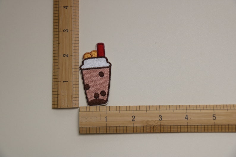 Może przedstawiać: Haftowana naszywka przedstawiająca brązowy nap&oacute;j bubble tea z białym obramowaniem, brązowymi perełkami boba i czerwoną słomką. Naszywka ma około 3,8 cm wysokości i 2,5 cm szerokości. Naszywka znajduje się na jasnobeżowym tle.