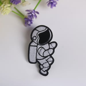 Könnte beinhalten: Ein gestickter Bügelaufkleber mit einem Astronauten in einem weißen und schwarzen Raumanzug.