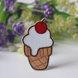 May include: An embroidered ice cream cone patch in white and brown with a red cherry on top.