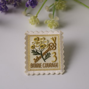 Pode incluir: Um remendo de tecido branco com um desenho bordado em ouro de uma flor e uma chave. O texto "BONNE COURAGE" também está bordado em ouro.