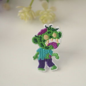 May include: Green and purple embroidered zombie patch with a white background. The zombie has a toothy grin and is wearing a blue shirt and purple pants.