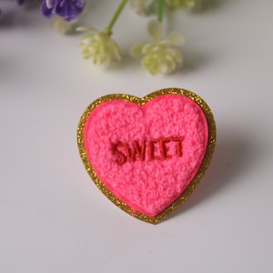 May include: A pink heart-shaped candy with a gold glitter border and the word "SWEET" embroidered in red.