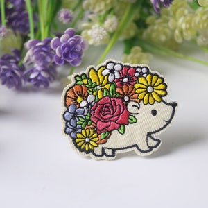 Könnte beinhalten: Gestickter Aufnäher mit einem lächelnden Igel mit einem bunten Blumenkranz. Die Blumen sind rote Rosen, gelbe Gänseblümchen und orangefarbene Blüten. Der Aufnäher hat einen cremefarbenen Hintergrund und eine schwarze Umrandung.
