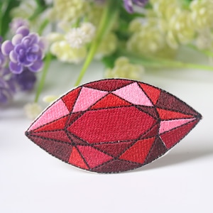 May include: An embroidered patch shaped like a gemstone, featuring red and pink colours. The patch is detailed with black stitching and has a faceted design.