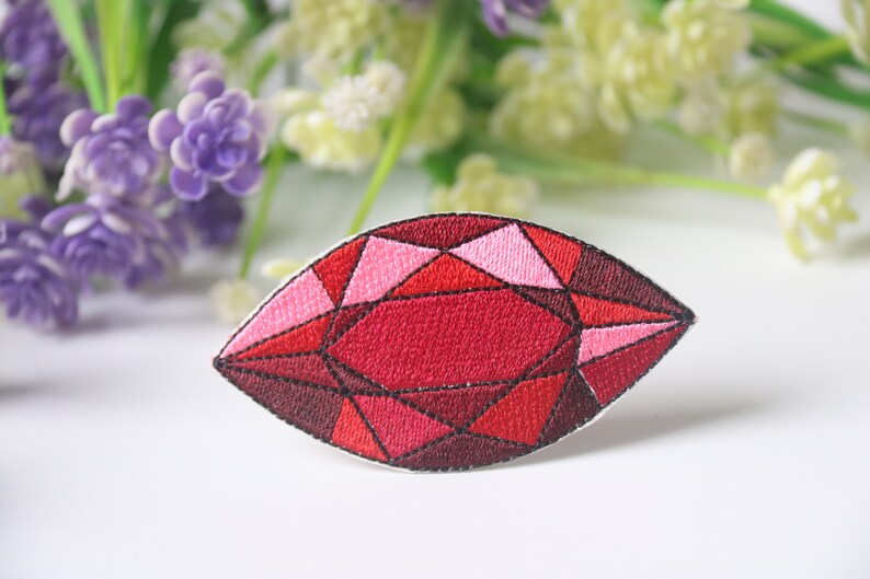 May include: An embroidered patch shaped like a gemstone, featuring red and pink colours. The patch is detailed with black stitching and has a faceted design.