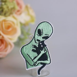 May include: Green alien embroidered patch with black outline holding a black cat with white eyes. The alien has large eyes and a small mouth.