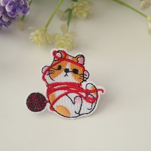 May include: Embroidered iron-on patch of a white cat with orange and brown markings tangled in red yarn with a red yarn ball.