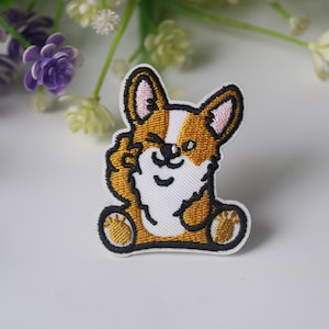 Puede incluir: Parche bordado para planchar con un perro corgi de dibujos animados con una expresión de guiño, sentado con las patas cruzadas. El corgi es marrón y blanco con contornos negros.