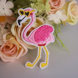 May include: Pink flamingo with white outline and yellow legs wearing heart-shaped sunglasses. The flamingo is embroidered on a white fabric.