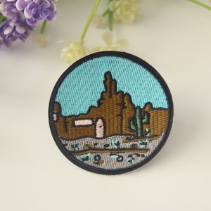 May include: Embroidered patch featuring a desert landscape with brown rock formations, a green cactus, and a light blue sky. The patch is circular with a black border. The design includes a building with a pink door.