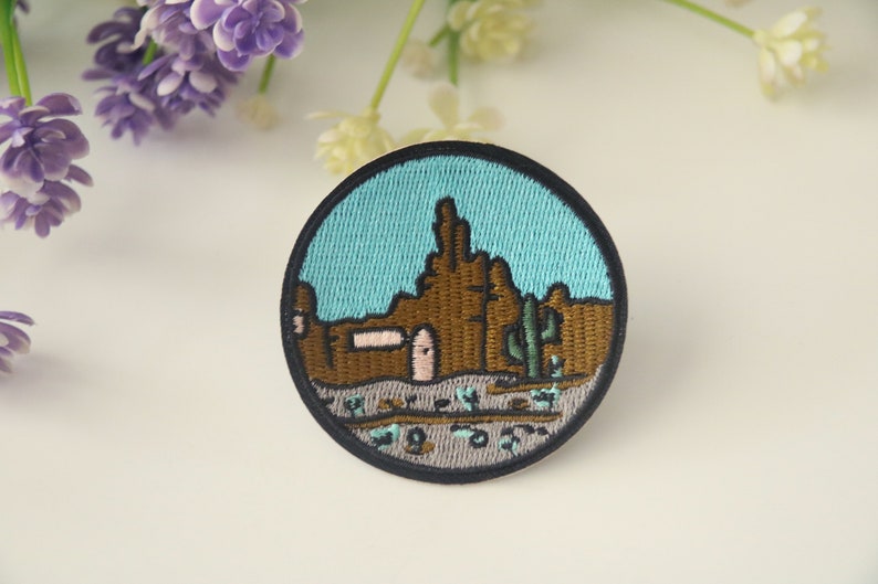 Peut inclure: &Eacute;cusson brod&eacute; circulaire repr&eacute;sentant un paysage d&eacute;sertique. Il pr&eacute;sente des formations rocheuses marron, un cactus vert et un ciel bleu clair, le tout bord&eacute; de noir. Un b&acirc;timent avec une porte rose est &eacute;galement repr&eacute;sent&eacute;.