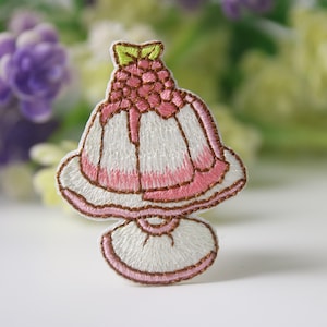 May include: A pink and white embroidered patch of a dessert with a raspberry on top. The dessert is on a white stand with a brown trim.