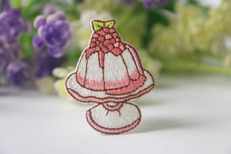Peut inclure: Un patch brod&eacute; repr&eacute;sentant un dessert rose et blanc avec une framboise sur le dessus. Le dessert est sur un support blanc avec une bordure brune.