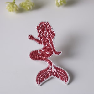 May include: A pink embroidered iron-on patch of a mermaid with a long flowing tail.