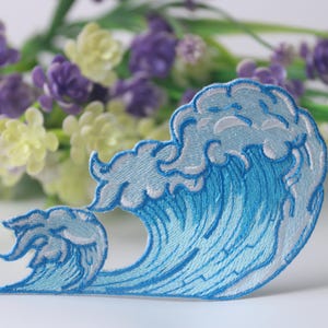 Può includere: Un toppa ricamata a forma di onda blu e bianca. L'onda è stilizzata e ha una cresta spumosa. La toppa è perfetta per aggiungere un tocco nautico a vestiti o accessori.