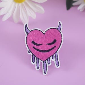 May include: Embroidered patch featuring a pink heart with closed eyes, devil horns, and dripping purple accents. The patch has a white border and is set against a light purple background. Daisies are in the background.