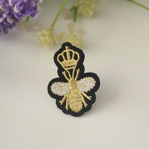 Może przedstawiać: Złota haftowana pszczoła z koroną na głowie, pszczoła znajduje się na czarnym filcowym plastrze.