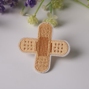May include: A beige embroidered iron-on patch shaped like a bandage with a white outline.