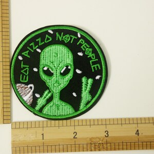 May include: Green alien with large eyes and a peace sign, embroidered on a black patch. The text around the alien reads "Eat Pizza Not People".