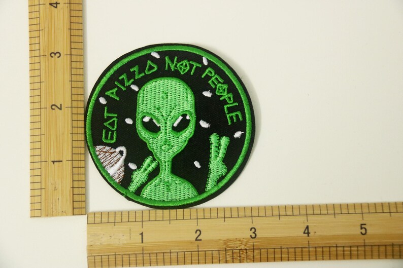 May include: Green alien with large eyes and a peace sign, embroidered on a black patch. The text around the alien reads "Eat Pizza Not People".