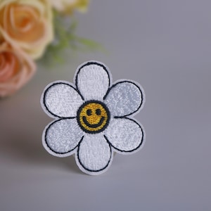 Puede incluir: Parche de flores blancas bordadas con una cara sonriente amarilla en el centro.