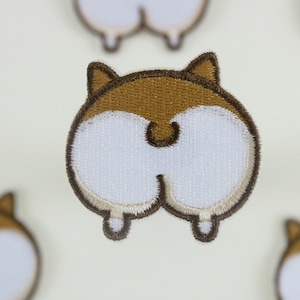 Könnte beinhalten: Brauner und weißer gestickter Aufnäher mit dem Hinterteil eines Hundes mit braunem Schwanz und Ohren.