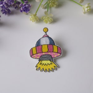 May include: Embroidered iron-on patch of a pink and yellow cartoon spaceship with a yellow flame.
