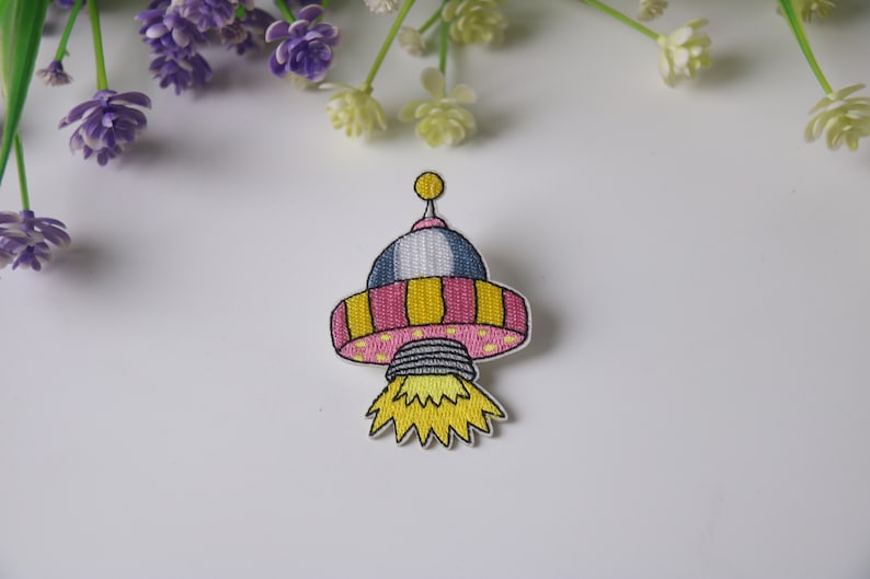 May include: Embroidered iron-on patch of a pink and yellow cartoon spaceship with a yellow flame.