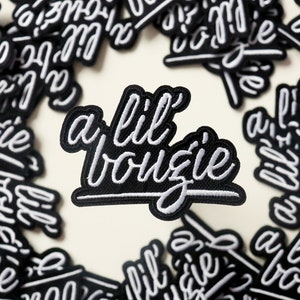 May include: A black and white embroidered patch with the text "a lil bougie" in a cursive font.