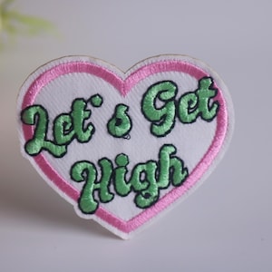 May include: A white heart-shaped patch with a pink outline. The patch has green embroidered text that reads "Let's Get High".