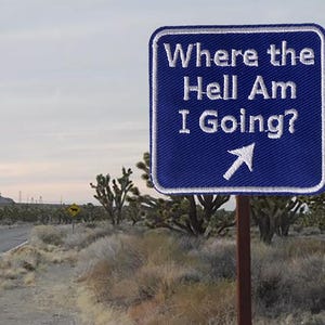 以下が含まれることがあります： 「Where the Hell Am I Going?」と白文字で書かれた青い看板。白い矢印が右を指している。看板は砂漠の風景の中で木の柱に取り付けられている。