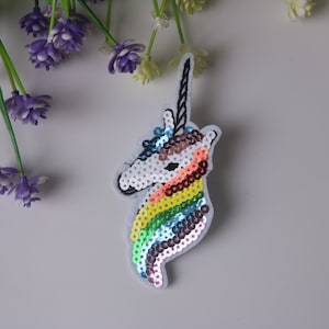 May include: A white unicorn head applique with a rainbow mane made of sequins. The unicorn has a silver horn and a white face.