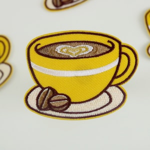 Op de afbeelding: Gele geborduurde strijkpatch van een koffiekopje met een hartvormig ontwerp in het schuim, op een schoteltje met een koffieboon.