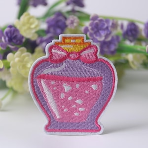 May include: Embroidered patch in the shape of a potion bottle. The patch features a pink potion with white accents inside a purple bottle. A pink bow and yellow top complete the design. The patch is outlined in white.
