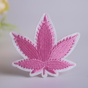 Pode incluir: Um patch de folha bordada rosa com uma borda branca. O design da folha é detalhado com costuras visíveis, e o patch é colocado em um fundo macio. O patch é um item decorativo.
