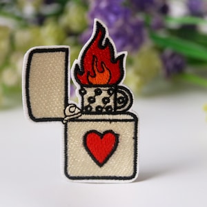 Puede incluir: Un parche bordado para planchar de un encendedor con un corazón rojo y una llama. El encendedor es blanco con contornos negros. La llama es roja y naranja.